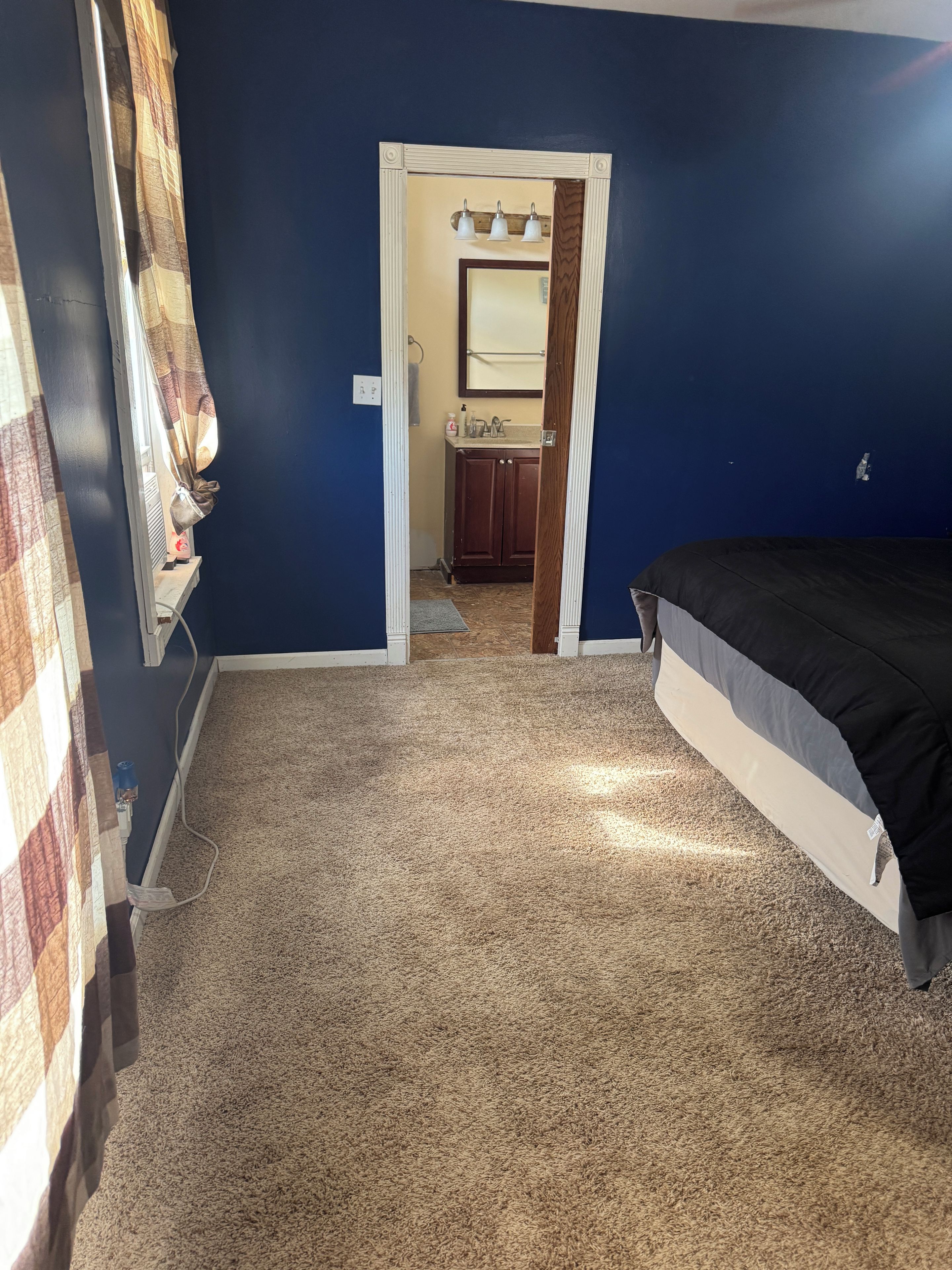 A bedroom with a bed, carpeted floor, and a bathroom visible through an open door.