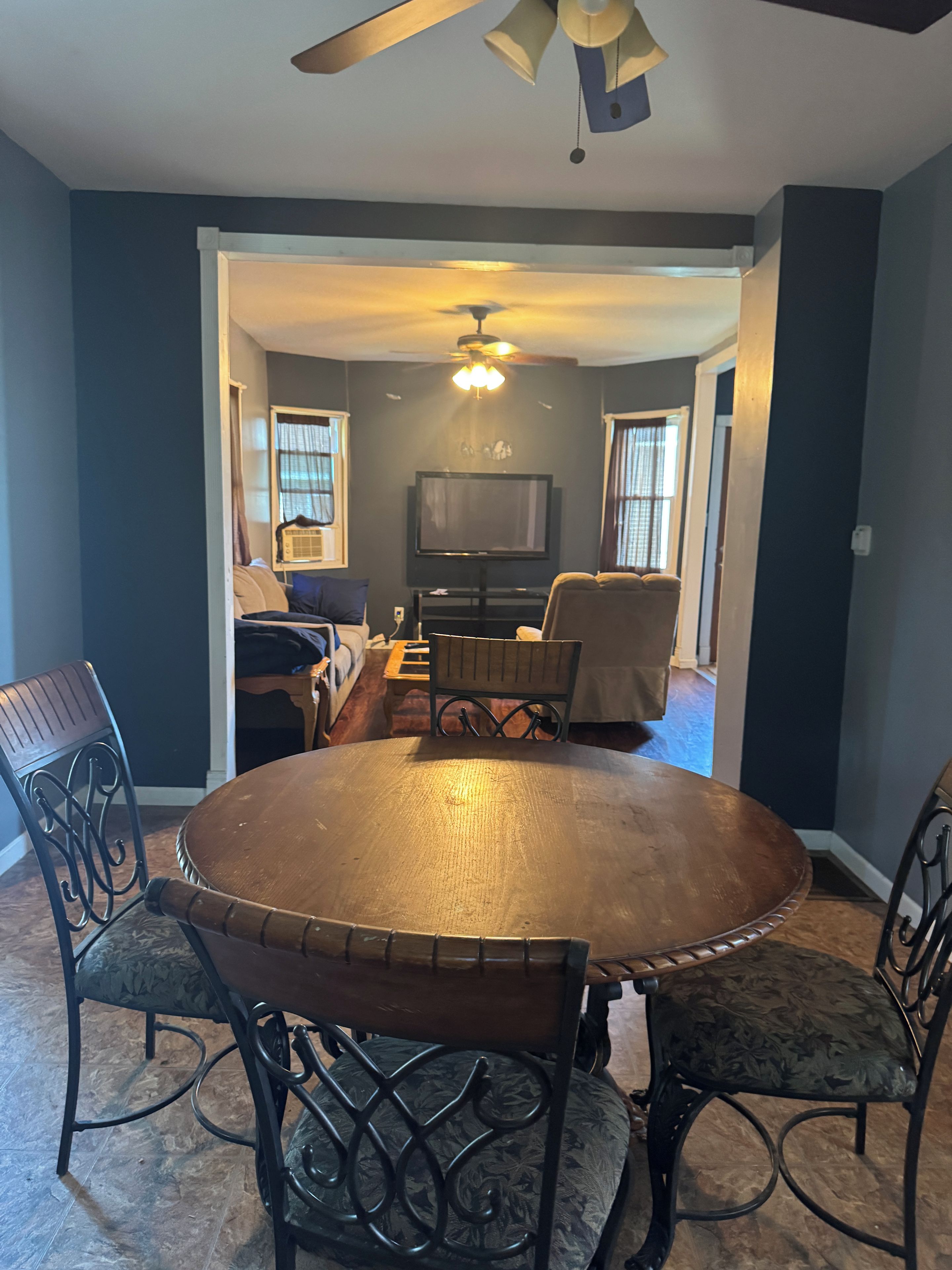 A dining area with a round table, four chairs, a ceiling fan, and a television in the background.