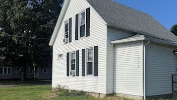 Une maison blanche à deux étages, dotée d’un toit à pignon et de volets noirs, entourée d’une pelouse verdoyante.