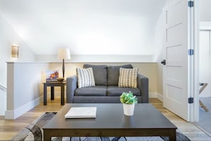 Una sala de estar con un sofá gris, dos almohadas estampadas, una mesita con una lámpara, un mueble bajo con una planta y un cuadro enmarcado.