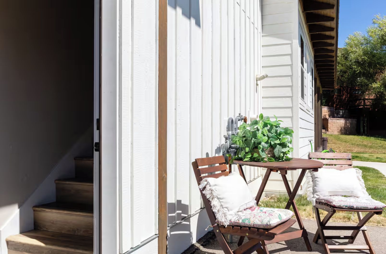 A wooden staircase leading to a white exterior wall with a small outdoor seating area featuring two wooden chairs and a table.
