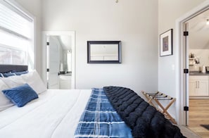 Un dormitorio con cama, un ventanal grande, un espejo, un cuadro enmarcado y vista a la cocina.