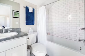 Un baño con inodoro blanco, un lavabo con encimera de mármol, ducha con cortina blanca y pared con azulejos.