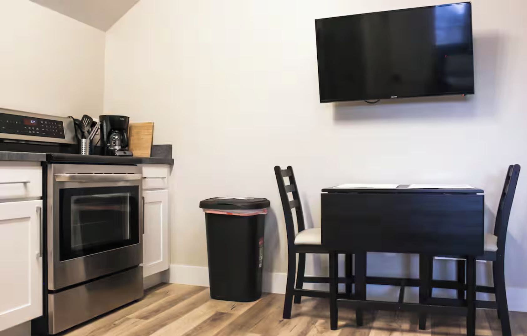 A modern kitchen with a built-in oven, a trash bin, a dining table with chairs, and a mounted television.
