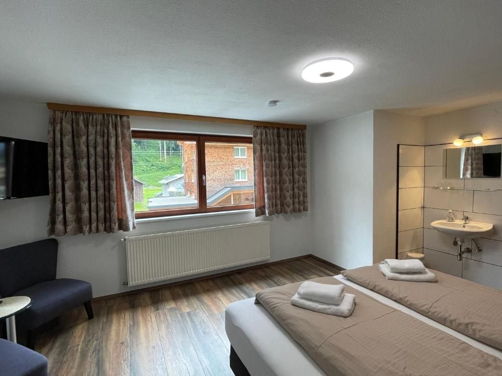 Habitación de hotel con cama, escritorio, silla, ventana con cortinas, radiador y baño con lavamanos y espejo.