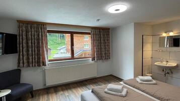 Habitación de hotel con cama, escritorio, silla, ventana con cortinas, radiador y baño con lavamanos y espejo.