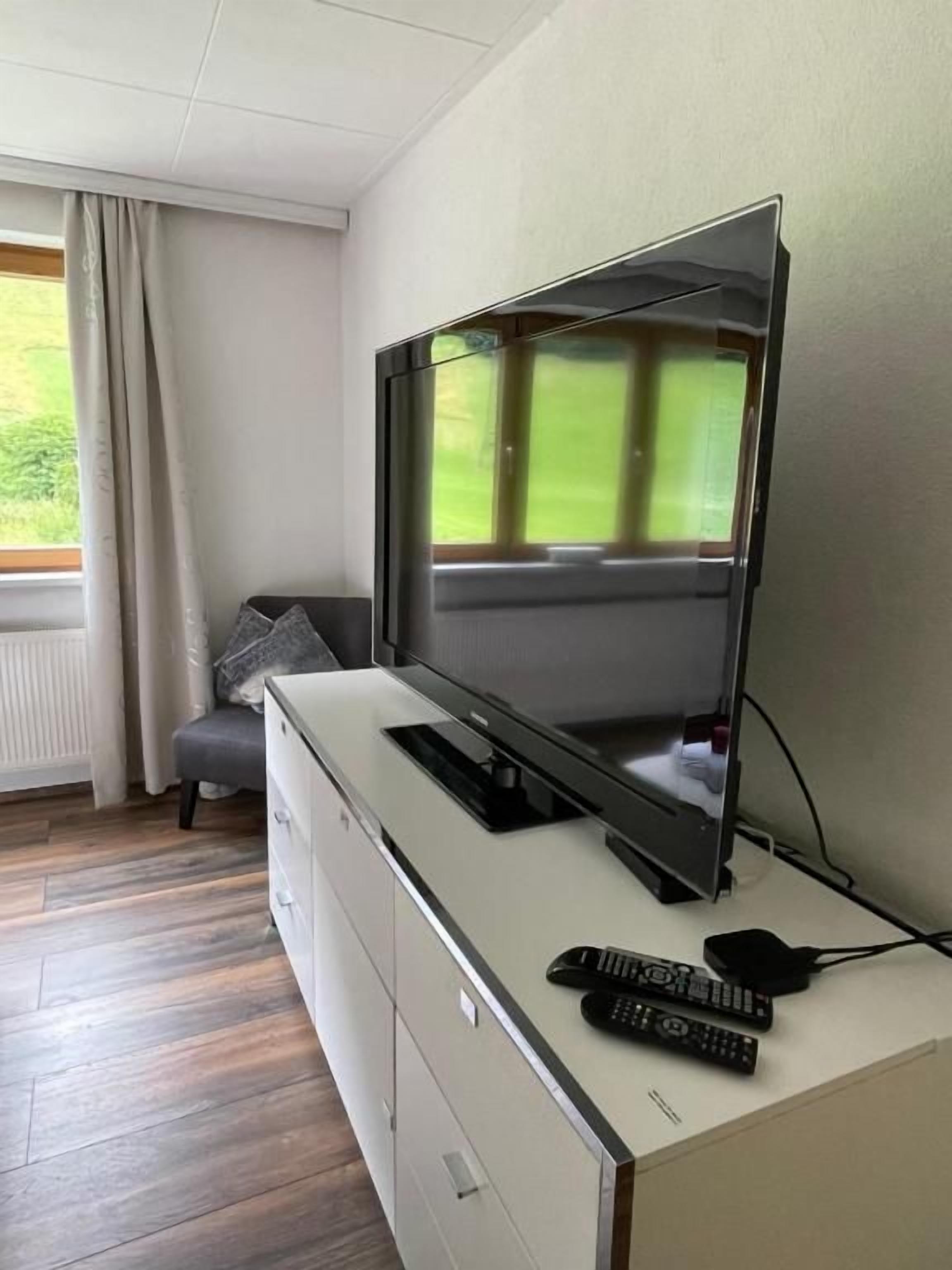 A modern living room with a flat-screen TV mounted on a cabinet, a remote control, and a window with curtains.