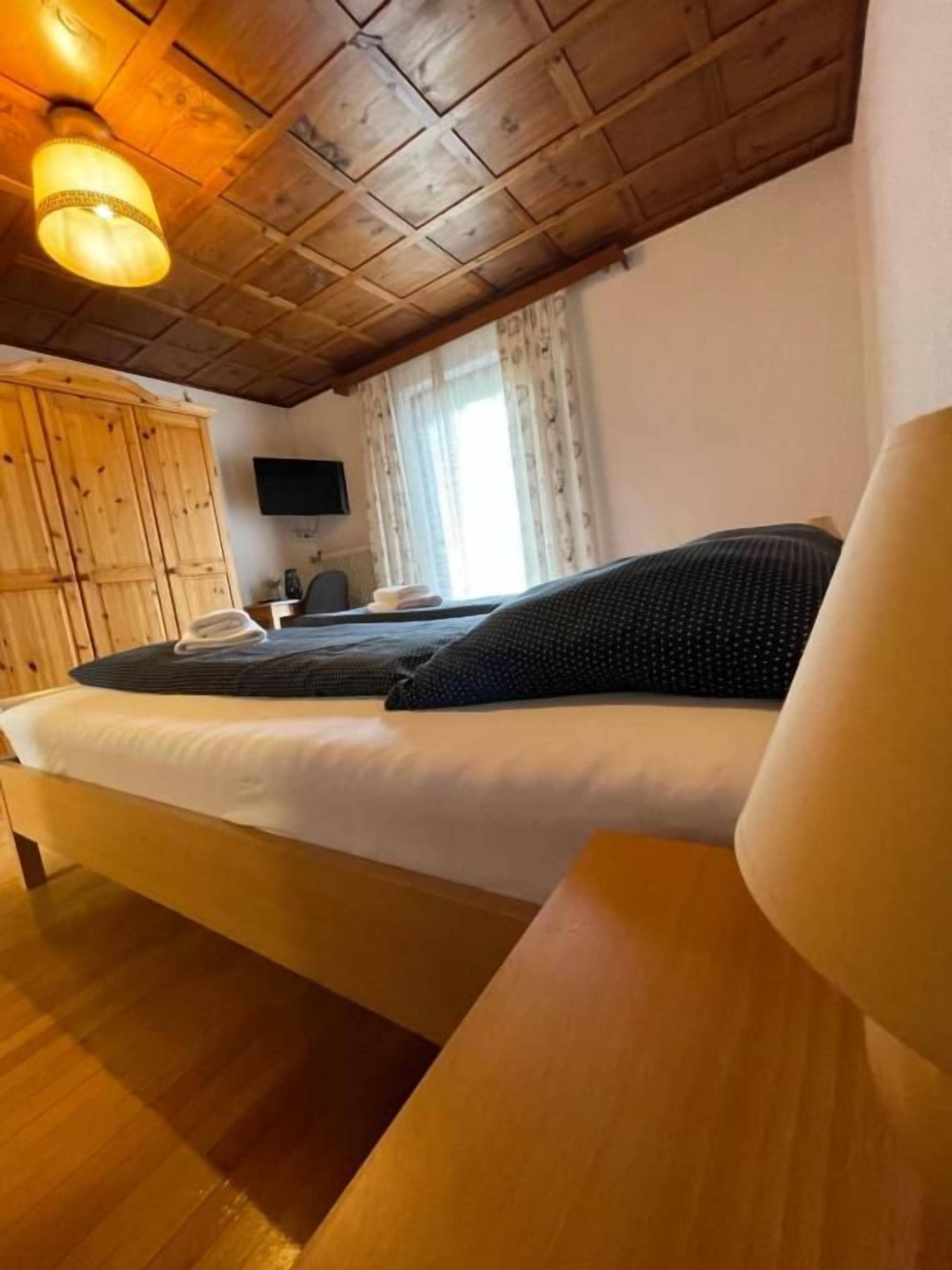 Un dormitorio con techo de madera, una cama con ropa oscura, una mesita de noche de madera y un televisor en la pared.