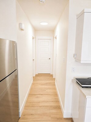 A narrow hallway with a wooden floor, white walls, a white door at the end, and a stainless steel refrigerator on the left. - Your Peaceful Cocoon at the Gates of Downtown (Riverview)