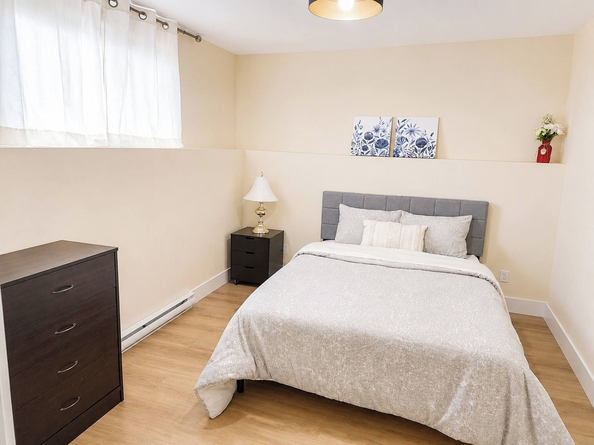 A bedroom with a bed, a dresser, a nightstand, a lamp, and artwork on the wall.