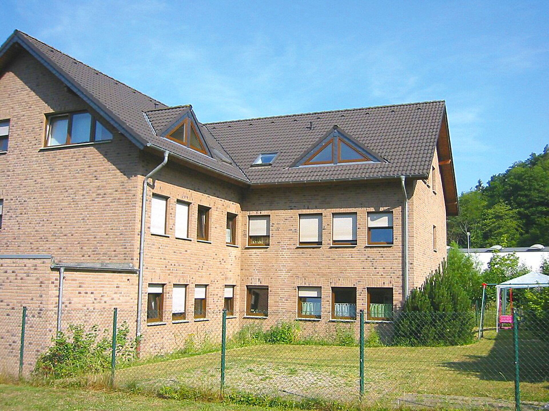 A multi-story brick building with a sloped roof, multiple windows, and a fenced outdoor area.