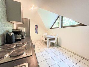Cozinha compacta com fogão, micro-ondas e cafeteira. Há uma mesa de jantar com cadeiras e uma janela com vista para o verde.