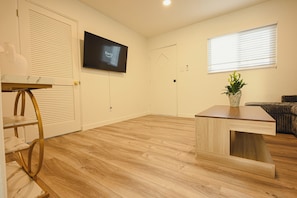 Un salón con piso de madera, un televisor de pantalla plana montado en la pared, una mesita pequeña y una planta en maceta.