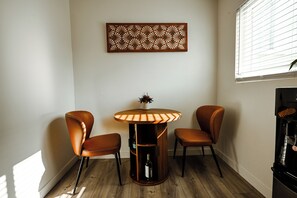 Een hoekzaal met een ronde tafel en twee stoelen, een decoratief kunstwerk aan de muur, en een raam met zonwering.