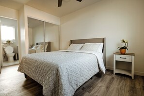 Een slaapkamer met een bed, een nachtkastje, een kamerplant en een badkamer zichtbaar door een open deur.