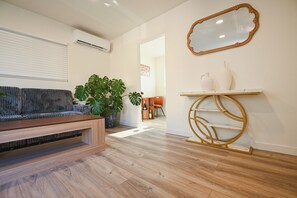 Een moderne woonkamer met een houten vloer, een grijze bank, een grote kamerplant en een decoratieve spiegel aan de muur.