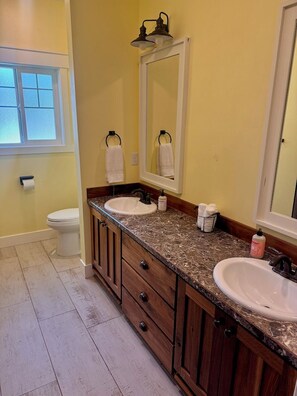 A bathroom with two sinks, wooden cabinets, a mirror, and a toilet. - The Gathering Place (Idaho Falls)