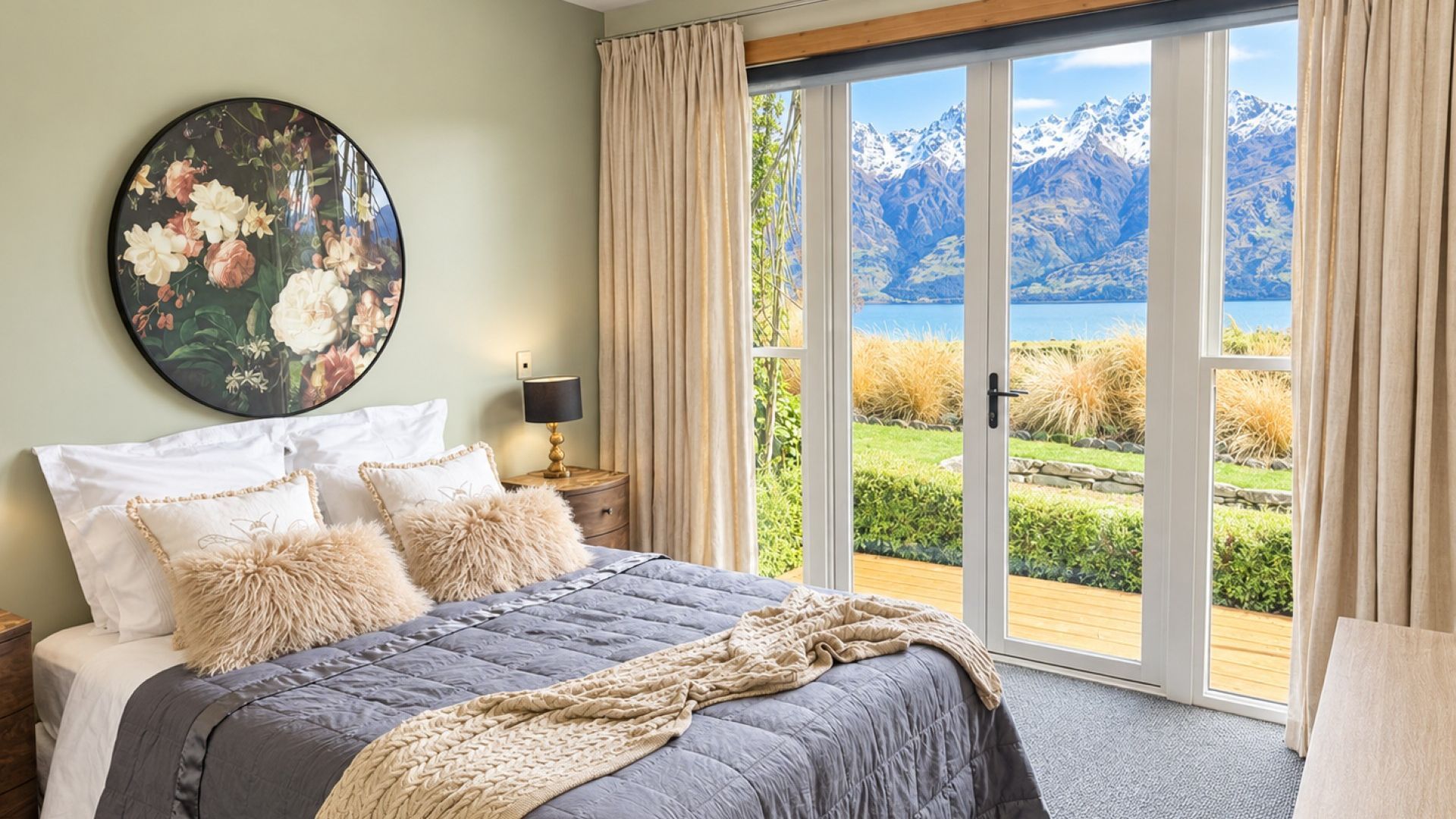 A bedroom with a bed, pillows, a nightstand, a lamp, a large window with curtains, and a view of mountains and a lake.