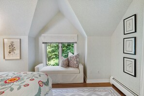 A bedroom with a sloped ceiling, a window with curtains, a bed with a floral bedspread, and framed botanical prints on the wall.