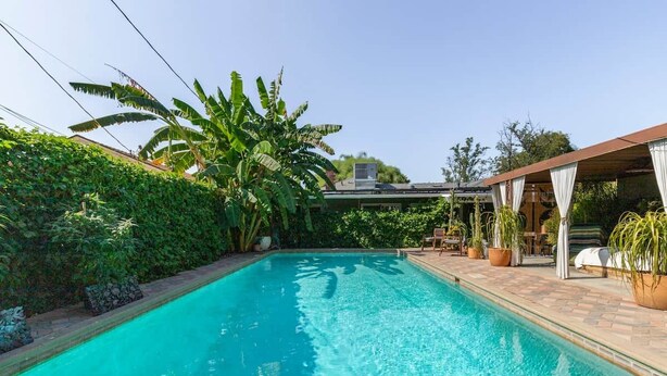 A swimming pool with a tiled deck, surrounded by lush greenery and a covered seating area. - Private 420 Pool & Spa Bungalow: Botanical Oasis (Los Angeles)