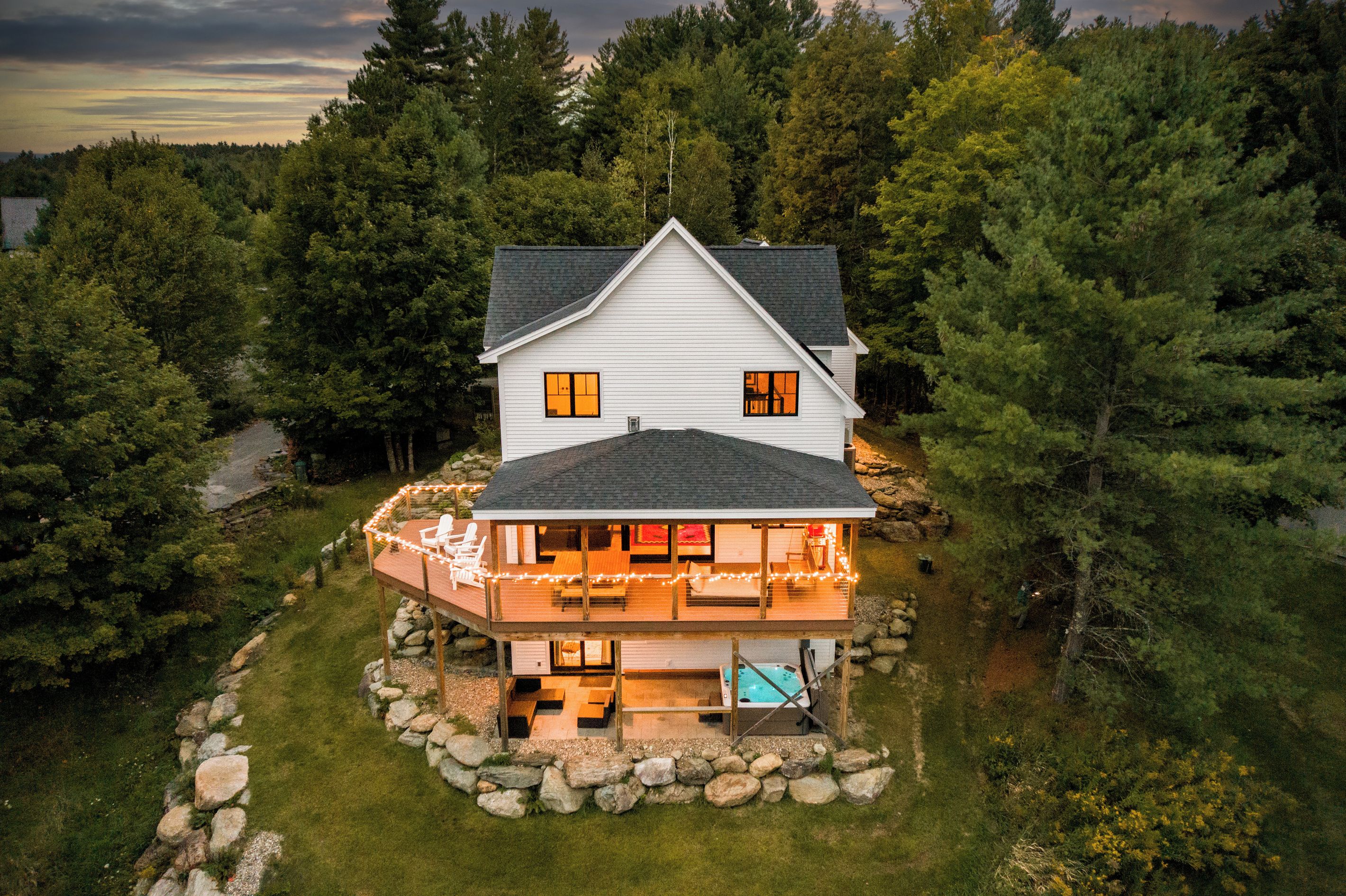 A two-story house with a deck and hot tub, surrounded by a stone wall and a forest.