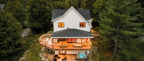 A two-story house with a deck and hot tub, surrounded by a stone wall and a forest.