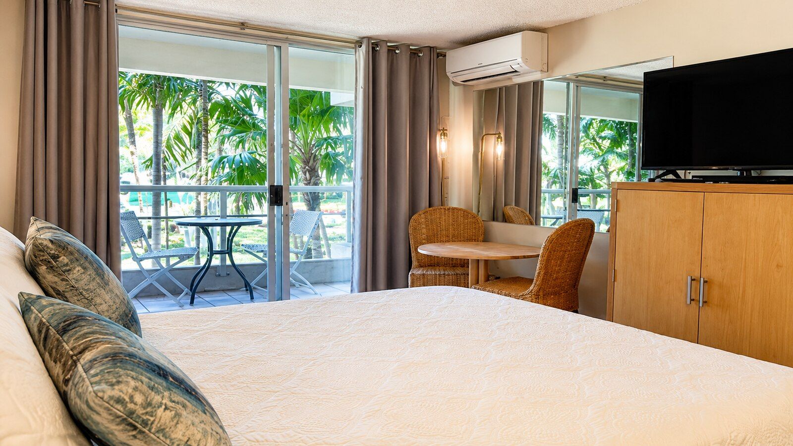 Habitación de hotel con una cama grande, televisor, armario de madera y balcón con mesa y sillas.