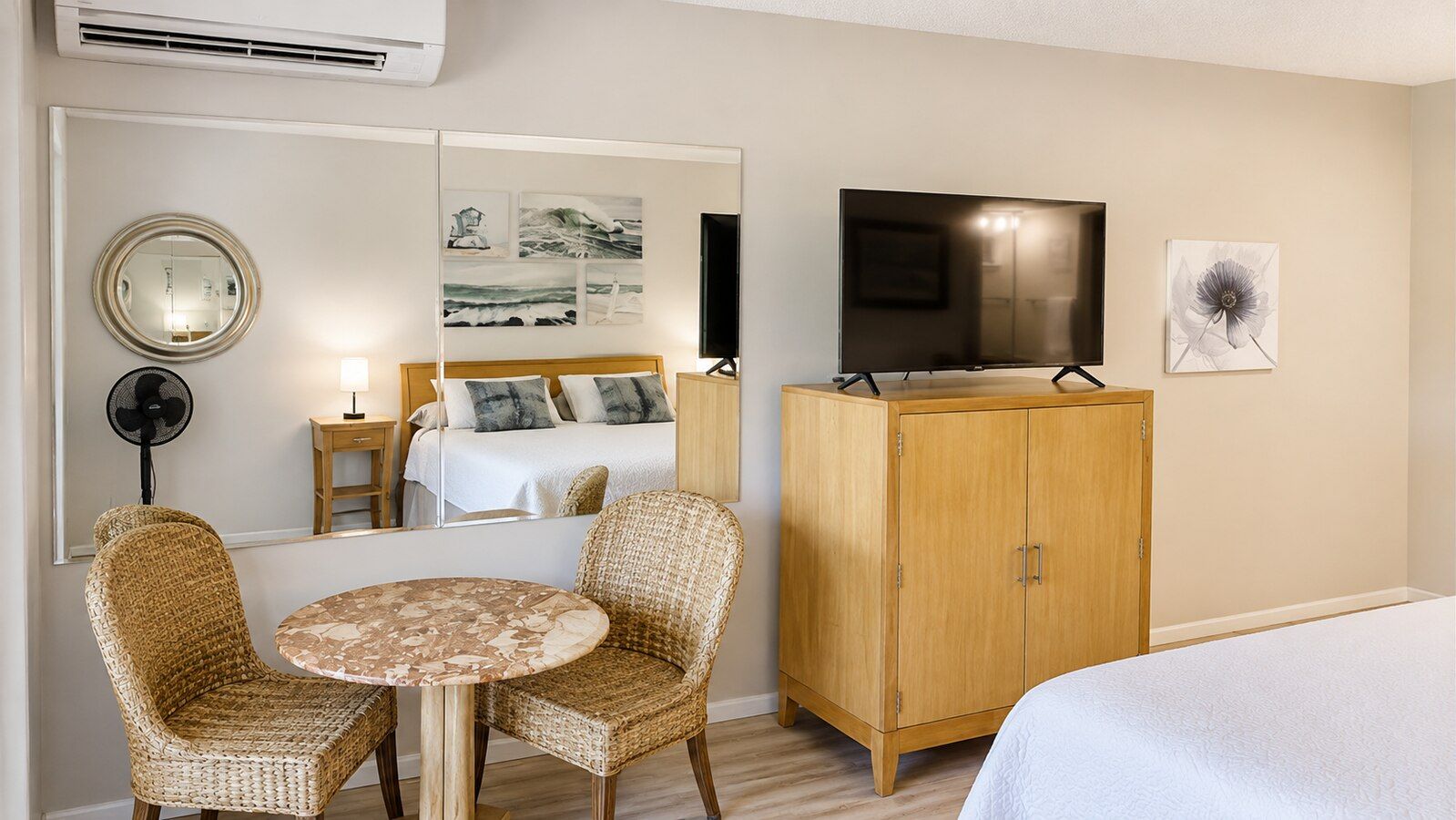 A hotel room with a bed, a wooden wardrobe, a television, a round table with two wicker chairs, a mirror, and artwork on the wall.