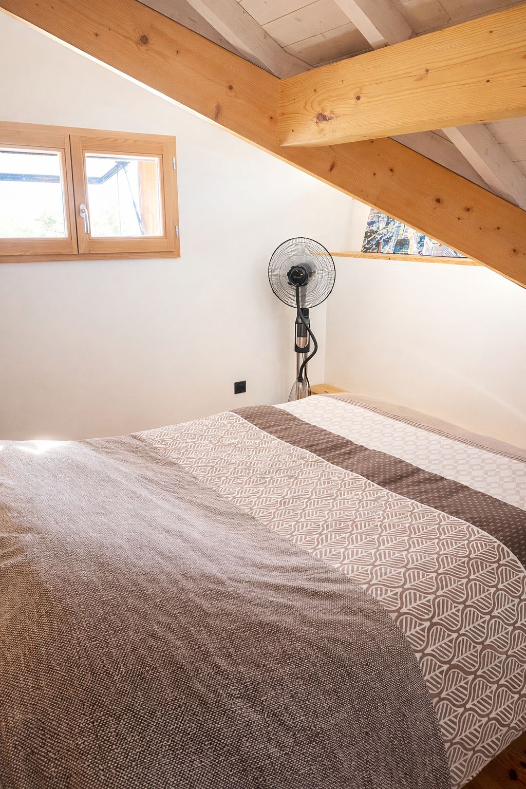 Ein Schlafzimmer mit schrägem Dach, einem Bett mit gemusterten Betttextilien, einem Standventilator und einem Fenster mit Blick.