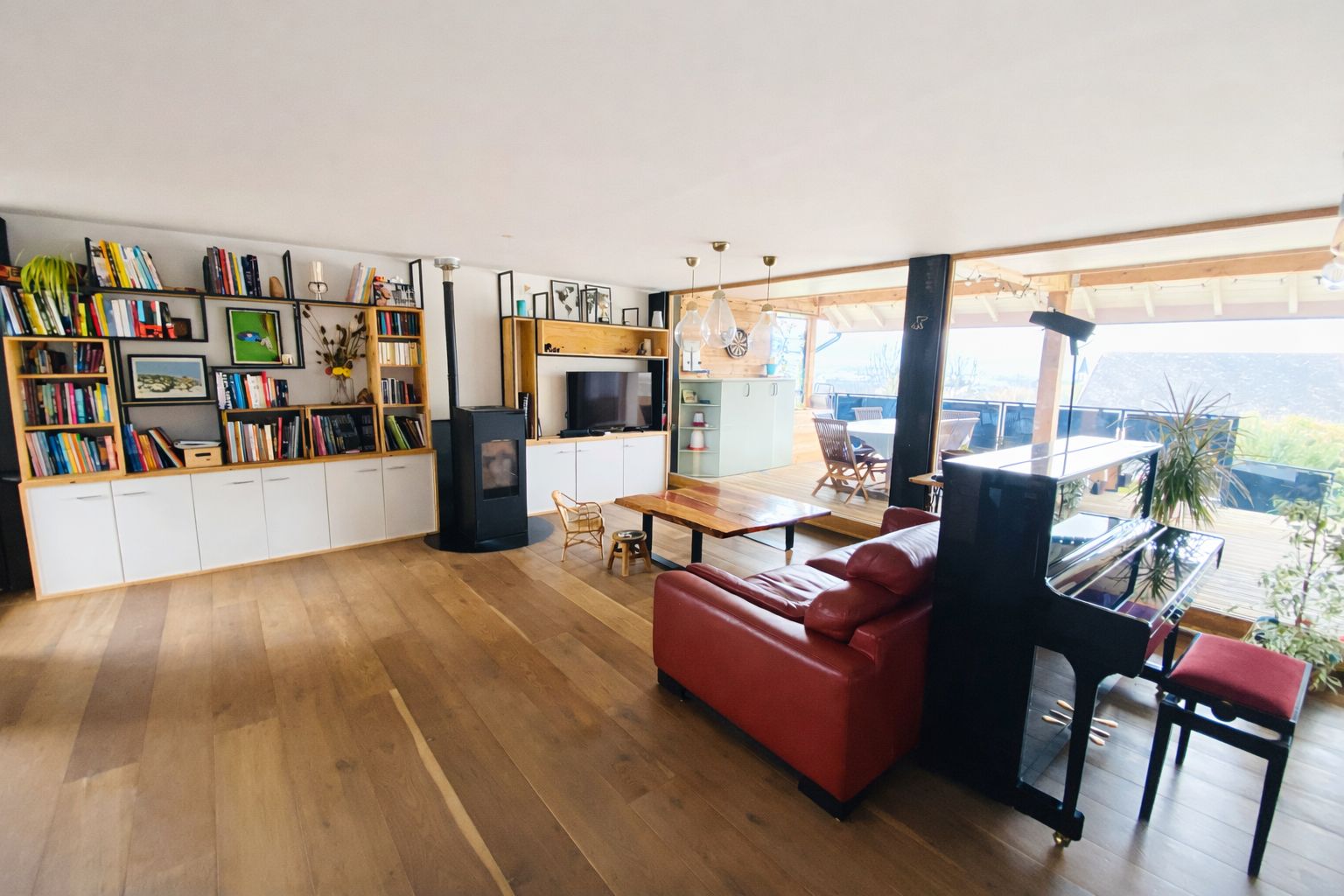 Un salon spacieux avec un canapé rouge, un plancher en bois, un coin repas comprenant une table et des chaises, une bibliothèque remplie de livres et une grande fenêtre offrant une vue dégagée.