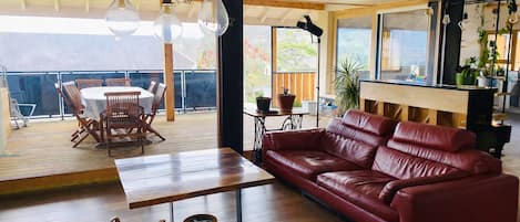 A spacious living area with a red leather sofa, wooden dining table, wicker chairs, and a balcony with outdoor seating.