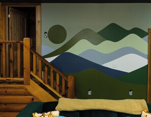Una habitación con escalera de madera, cama con manta verde y un mural en la pared que representa un paisaje con colinas y un sol.