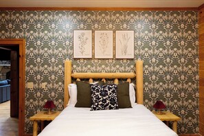 Un dormitorio con cabecera de madera, cama con ropa de cama blanca, dos mesitas de noche con lámparas y un papel tapiz floral.