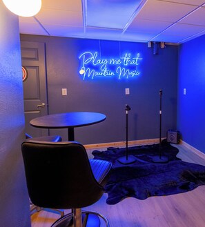 Una habitación con un letrero de neón que dice 'Play me that Mountain Music', una mesa redonda, una silla negra y un altavoz en el suelo.