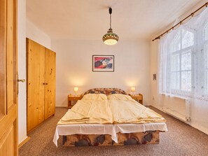 Une chambre avec une armoire en bois, un lit doté d’une tête de lit à motifs, deux lampes de chevet, un tableau encadré au mur et une fenêtre ornée de rideaux en dentelle.