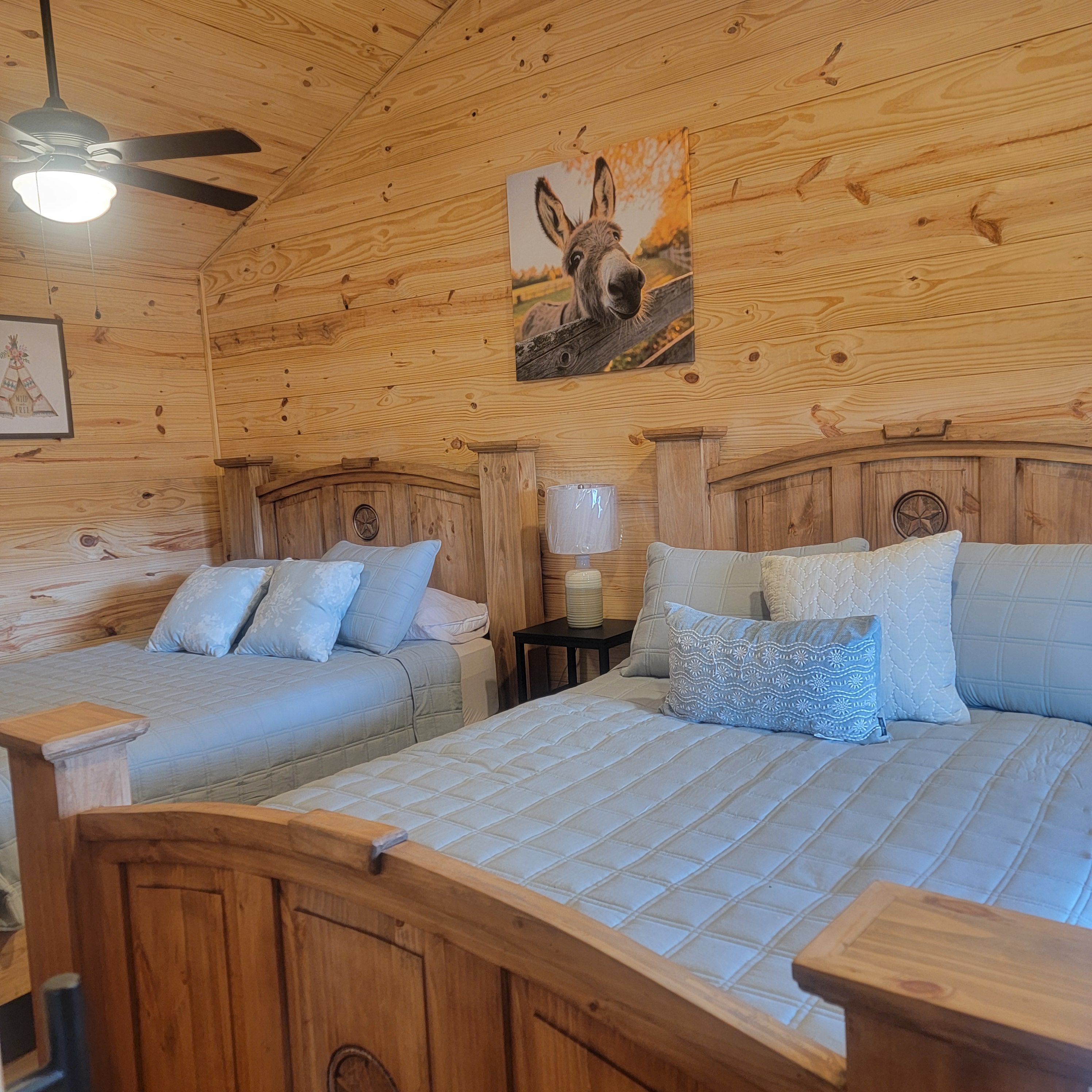 Una habitación tipo cabaña de madera con dos camas, un ventilador de techo y un cuadro colgado en la pared de un burro.