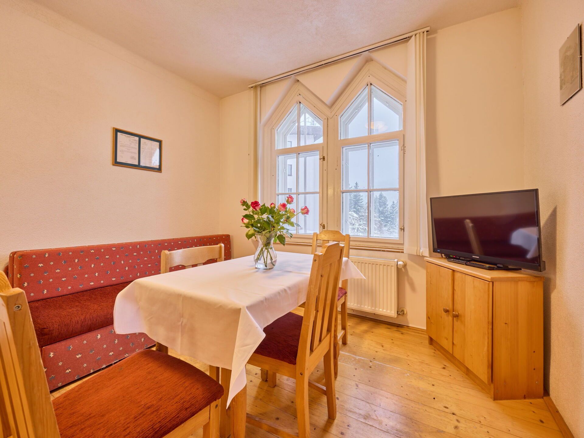 Une pièce chaleureuse avec une table en bois, un canapé rouge, un téléviseur posé sur un meuble en bois et une fenêtre donnant sur des arbres.