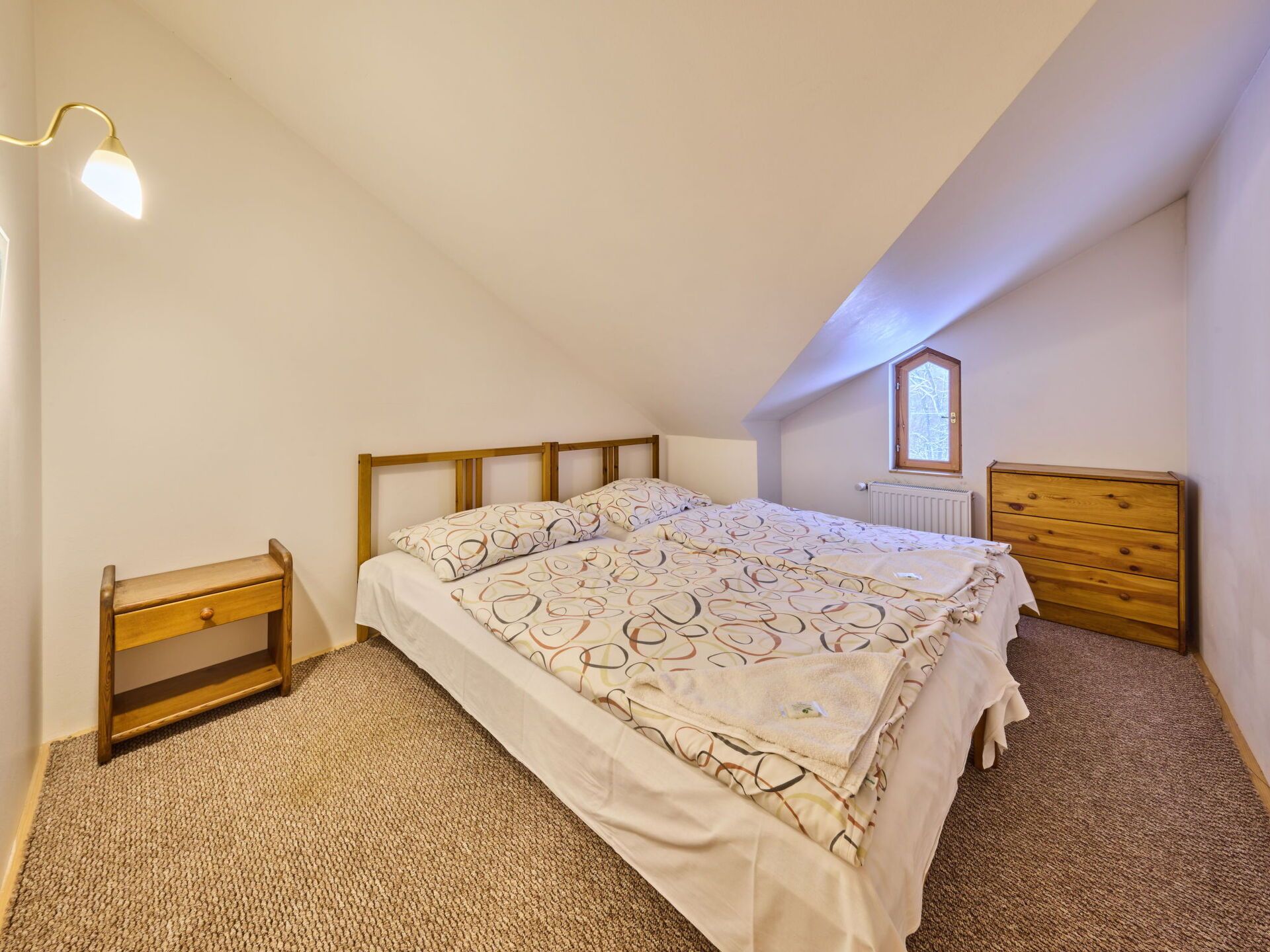 Une chambre avec un plafond en pente, un lit avec une literie à motifs, une table de chevet en bois, une commode et un radiateur.