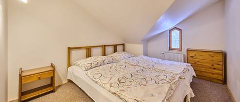 A bedroom with a sloped ceiling, a bed with patterned bedding, a wooden nightstand, a dresser, and a radiator.