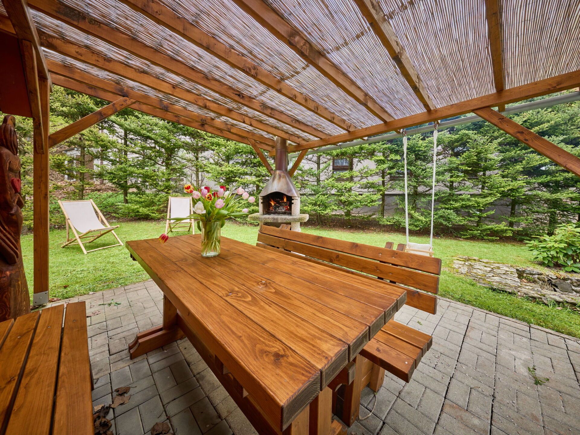 Une table en bois installée à l’extérieur, avec des bancs, un four à pizza en pierre et une chaise en bois.
