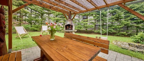 Une table en bois installée à l’extérieur, avec des bancs, un four à pizza en pierre et une chaise en bois.
