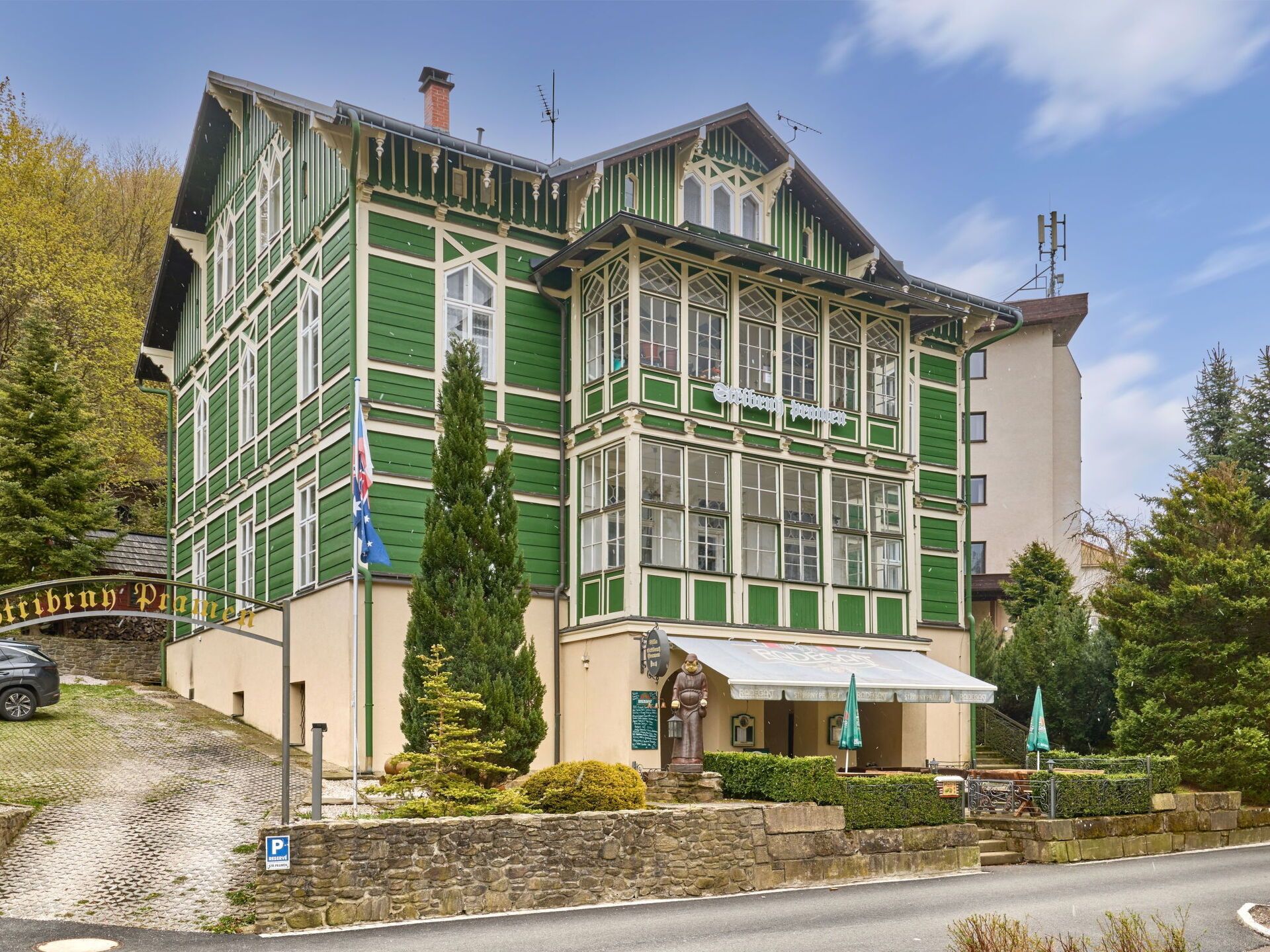 Un edificio tradicional con fachada verde, un muro de piedra en primer plano y acceso pavimentado.