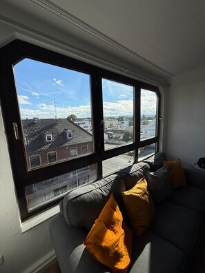 Una habitación con un ventanal grande, un sofá gris con cojines amarillos y vistas a edificios y un cielo despejado.