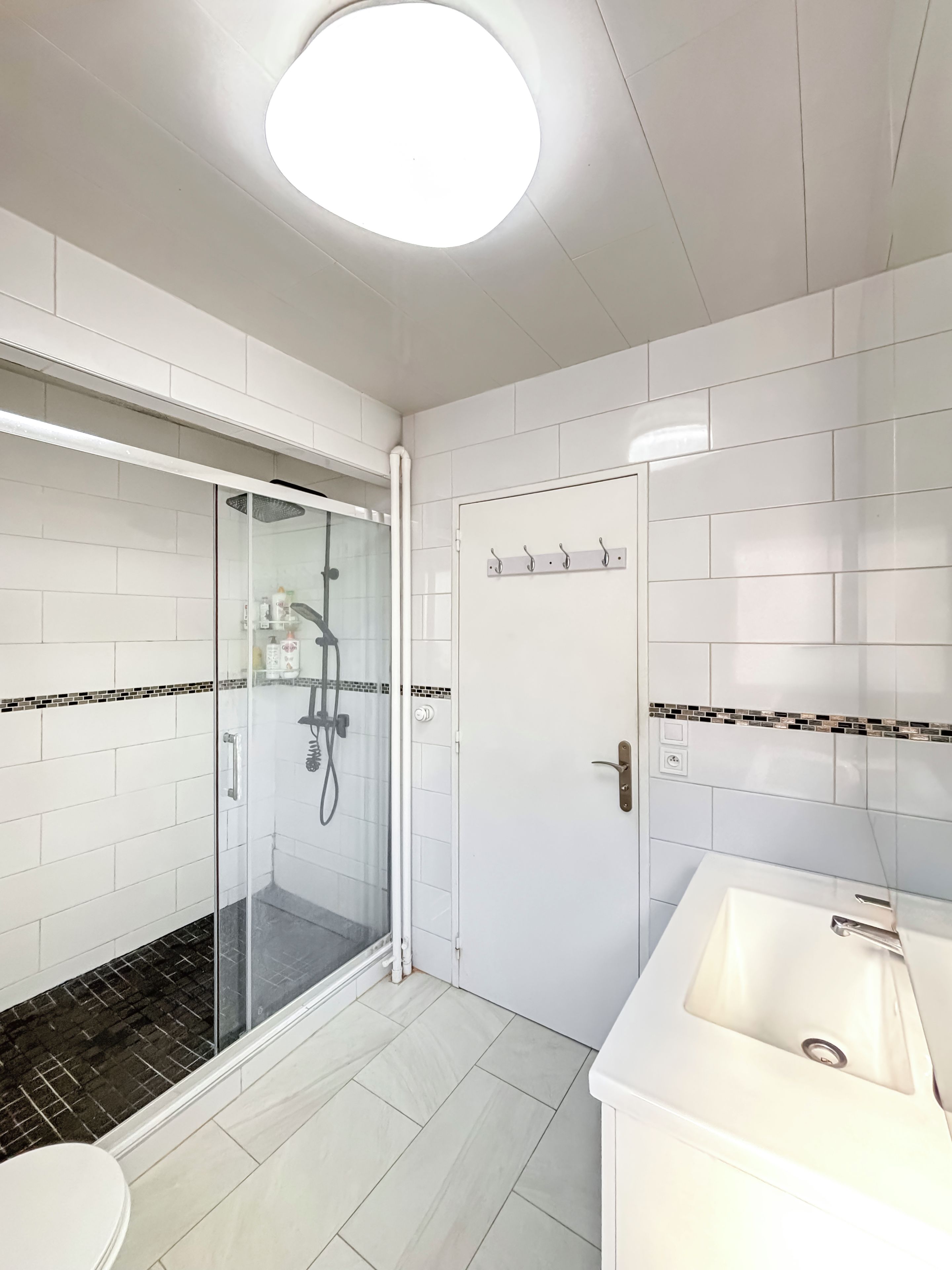 Een moderne badkamer met een douchecabine van glas, witte tegels, een wastafel en een deur met haken.