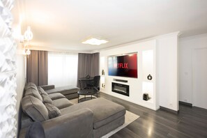 A modern living room with a large flat-screen TV mounted on the wall, a grey sectional sofa, a small round coffee table, and a fireplace below the TV. - The Art of Dreams - Near the Metro with Parking (Créteil)