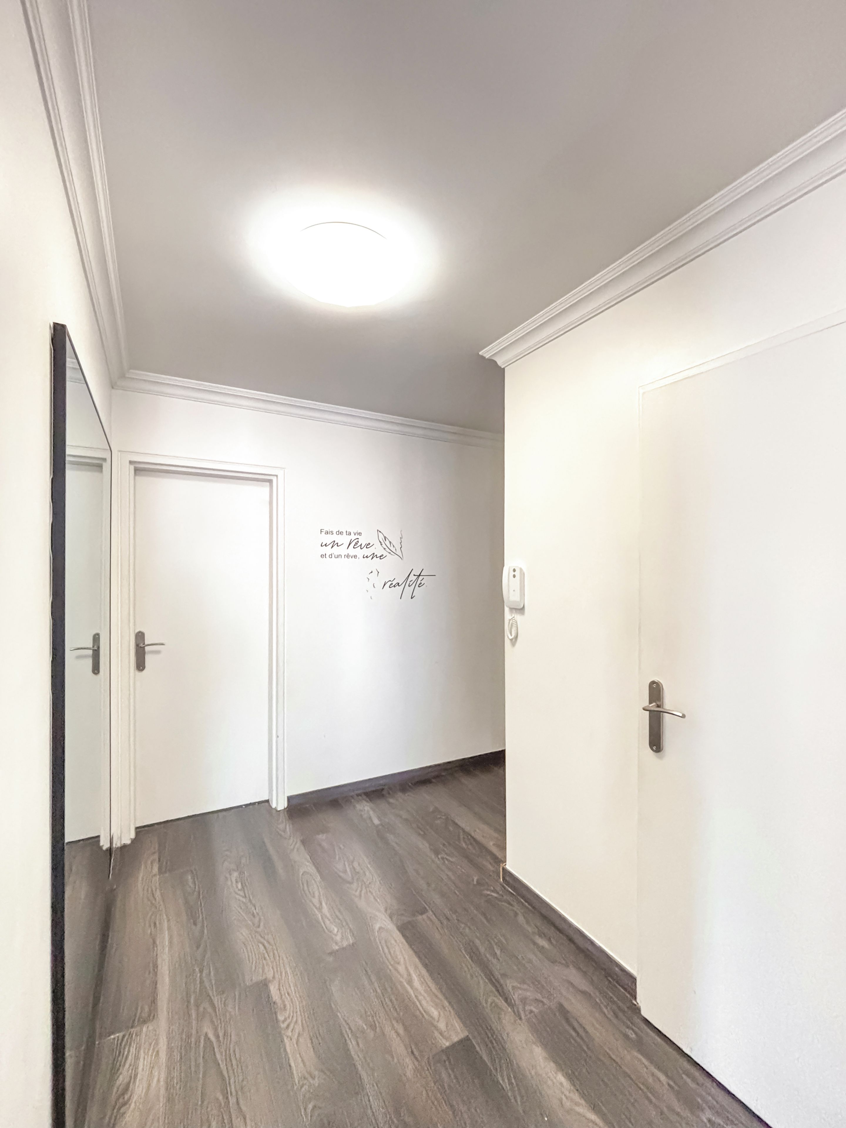 Un couloir doté d’un plancher en bois, de murs blancs et d’une porte ornée d’un motif décoratif.