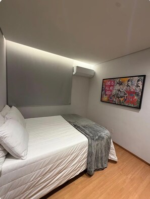 A bedroom with a bed, a gray blanket, a white pillow, a wall-mounted air conditioner, and a colorful abstract painting.