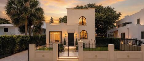 A modern two-story house with a symmetrical facade, a central entrance with steps, and a gated driveway.