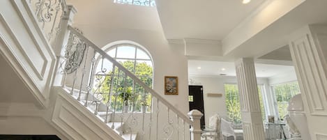 A grand staircase with ornate metal railings leading to a second floor, a chandelier, and a view of greenery outside.