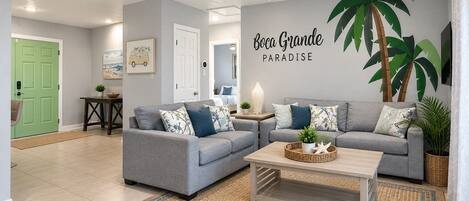 A living room with two grey sofas, a coffee table, a ceiling fan, and a wall with a large green palm tree mural.